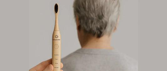 Hand holding a Crescent Nest bamboo electric toothbrush in focus, with an older adult blurred in the background, symbolizing the link between oral and brain health.