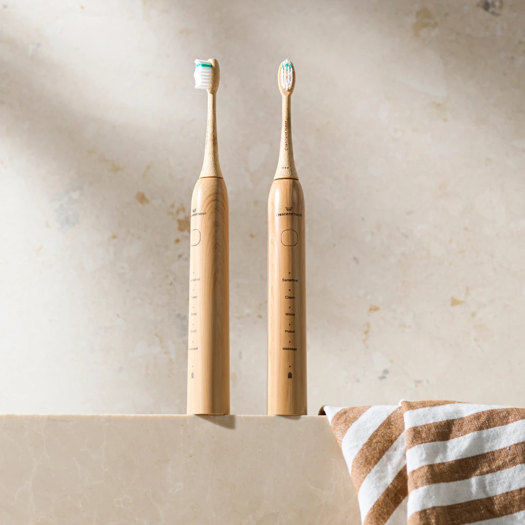 Two Crescent Nest bamboo electric toothbrushes standing upright in a bathroom setting with a striped towel beside them.