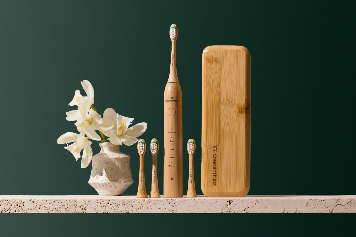 Crescent Nest bamboo electric toothbrush displayed with three replacement brush heads, a ceramic vase with white flowers, a glass of water, and a green cloth on a bathroom shelf.