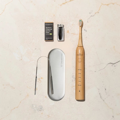 crescent nest Bamboo sonic toothbrush with a Stainless Steel Tongue Scraper, and bamboo dental floss on a beige surface