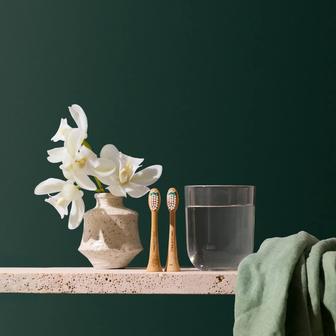 Bathroom counter with a vase of white flowers, toothbrushes, glass of water, and green towel against a dark green background.